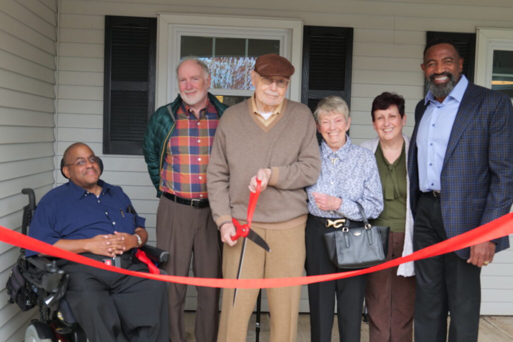A group of OTC Board members at the opening of the Robert Bankard Group Home. Robert Bankard is holding a large pair of scissors and is about to cut a red ribbon in front of the house.